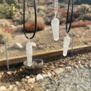 May include: Three silver wire wrapped clear quartz crystal pendants on different black and silver chains. The pendants are hanging against a blurred background of rocks and trees.