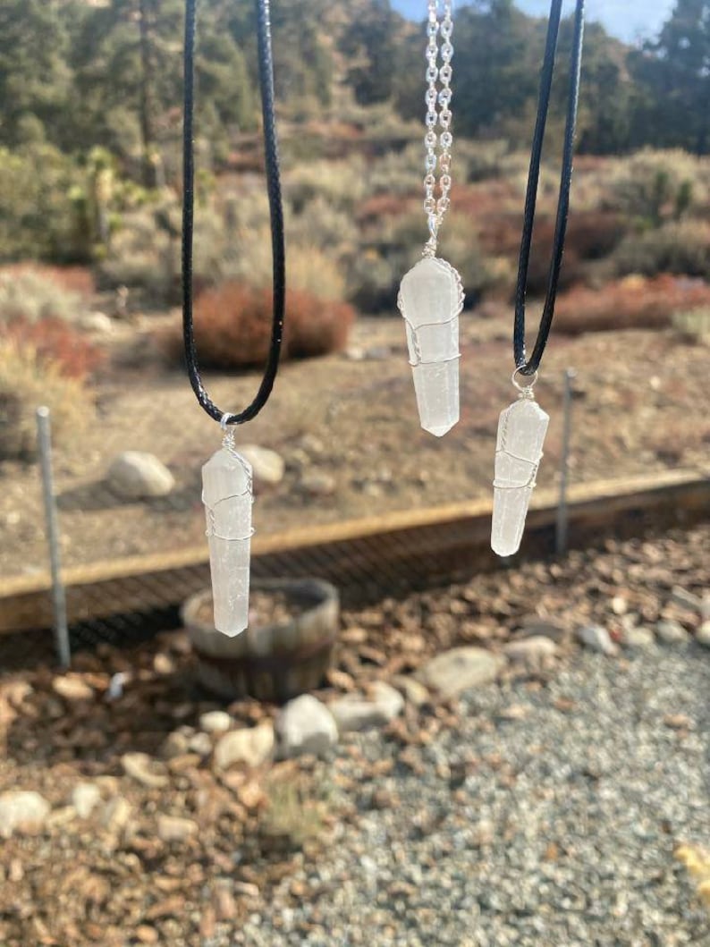 May include: Three silver wire wrapped clear quartz crystal pendants on different black and silver chains. The pendants are hanging against a blurred background of rocks and trees.