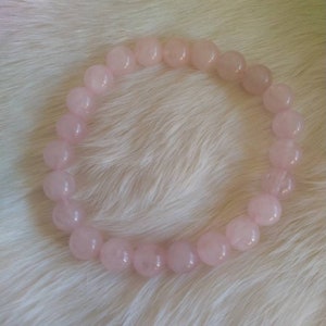 May include: A light pink rose quartz beaded bracelet. The bracelet is made of round, polished beads. The beads are strung together to form a circular shape. The bracelet is displayed on a white, fluffy surface.