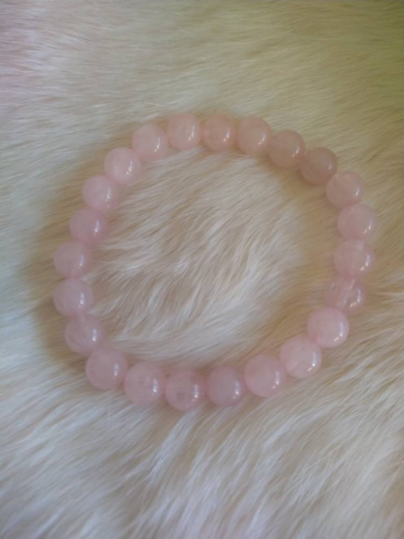 May include: A light pink rose quartz beaded bracelet. The bracelet is made of round, polished beads. The beads are strung together to form a circular shape. The bracelet is displayed on a white, fluffy surface.