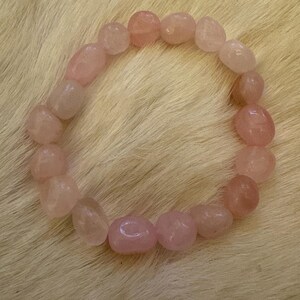 May include: A rose quartz bracelet composed of smooth, oval-shaped beads in varying shades of pink. The beads are strung together to form a circular shape, resting on a textured, light-colored surface. The bracelet is a piece of jewelry.