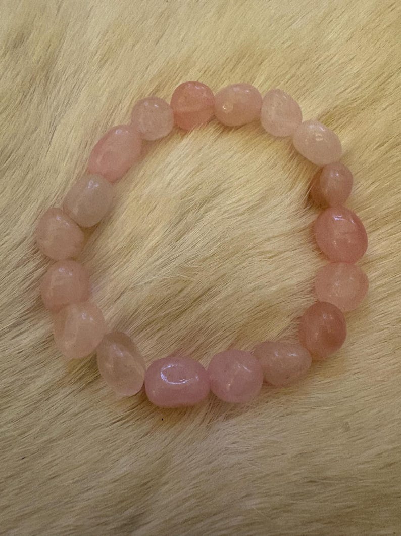 May include: A rose quartz bracelet composed of smooth, oval-shaped beads in varying shades of pink. The beads are strung together to form a circular shape, resting on a textured, light-colored surface. The bracelet is a piece of jewelry.