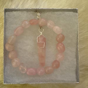 May include: A rose quartz necklace and bracelet set displayed in a silver box. The bracelet is made of rounded pink stones. The necklace features a rose quartz pendant wrapped in silver wire, suspended from a delicate chain. The set is presented on a white, textured surface.