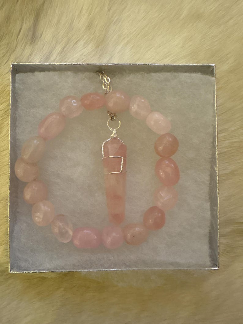 May include: A rose quartz necklace and bracelet set displayed in a silver box. The bracelet is made of rounded pink stones. The necklace features a rose quartz pendant wrapped in silver wire, suspended from a delicate chain. The set is presented on a white, textured surface.