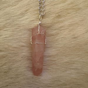 May include: A rose quartz crystal pendant necklace. The pink crystal is wrapped in silver-colored wire and hangs from a silver chain. The pendant is a long, hexagonal shape. The background is a light brown, furry texture.