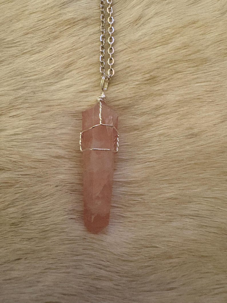 May include: A rose quartz crystal pendant necklace. The pink crystal is wrapped in silver-colored wire and hangs from a silver chain. The pendant is a long, hexagonal shape. The background is a light brown, furry texture.
