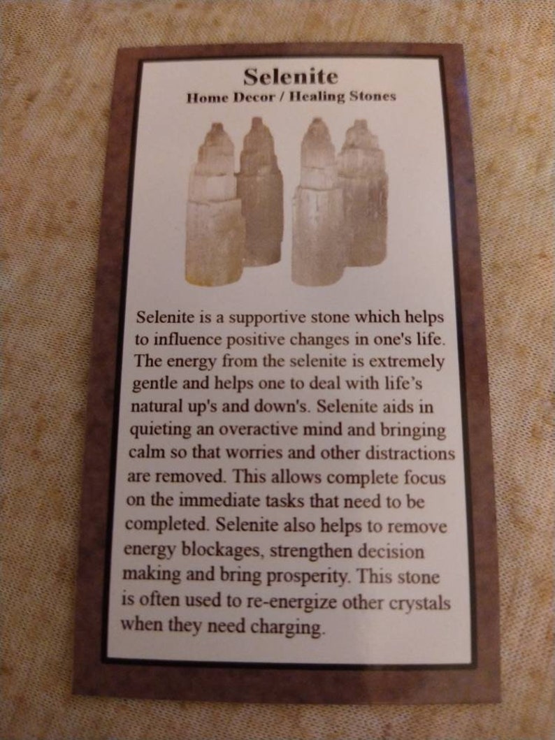 May include: A rectangular informational card with a brown border featuring a graphic of four off-white selenite stone towers. The text reads Selenite Home Decor / Healing Stones followed by a paragraph describing the properties of the stone.