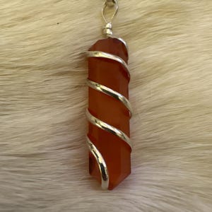 Op de afbeelding: Een hangerketting met een gepolijst, oranjerood kristal gewikkeld in zilverkleurig draad. Het kristal heeft een zeshoekige vorm. De ketting is zilverkleurig.