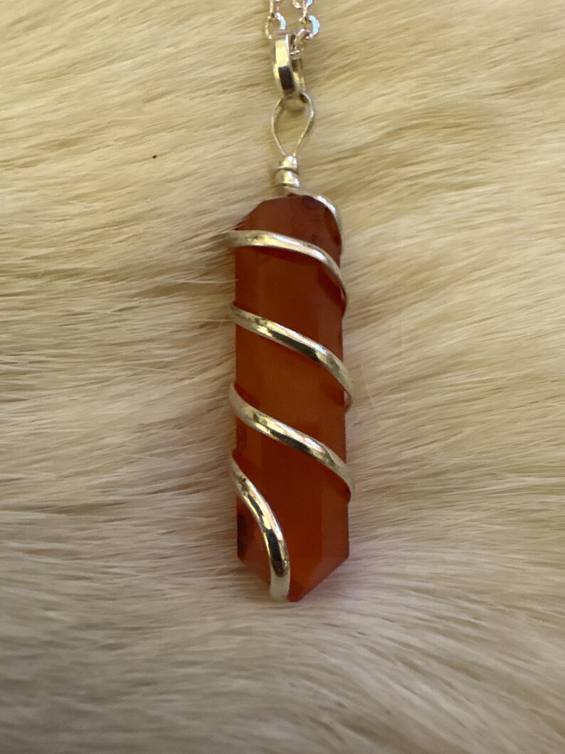 Op de afbeelding: Een hangerketting met een gepolijst, oranjerood kristal gewikkeld in zilverkleurig draad. Het kristal heeft een zeshoekige vorm. De ketting is zilverkleurig.