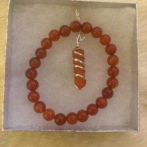 Op de afbeelding: Een carneliaan kralenarmband en hangerketting set. De armband heeft ronde, oranje-rode kralen. De hanger heeft een cilindrische carneliaan steen gewikkeld in zilverkleurig draad. De sieraden worden gepresenteerd in een kleine, zilveren geschenkdoos.