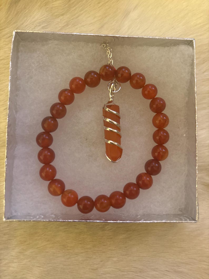 Op de afbeelding: Een carneliaan kralenarmband en hangerketting set. De armband heeft ronde, oranje-rode kralen. De hanger heeft een cilindrische carneliaan steen gewikkeld in zilverkleurig draad. De sieraden worden gepresenteerd in een kleine, zilveren geschenkdoos.