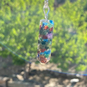 May include: A silver chain necklace with a clear crystal pendant filled with colorful gemstones. The pendant is shaped like a long, thin rectangle with a pointed end.