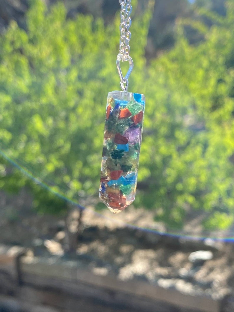 May include: A silver chain necklace with a clear crystal pendant filled with colorful gemstones. The pendant is shaped like a long, thin rectangle with a pointed end.