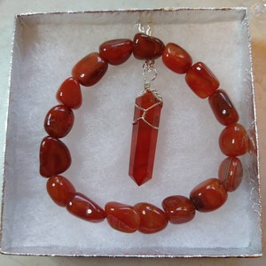 Op de afbeelding: Een carneliaan edelsteen armband en hanger set. De armband is gemaakt van gepolijste, rood-oranje stenen. De hanger is een bijpassende carneliaan kristal gewikkeld in zilverdraad. De set wordt gepresenteerd in een witte geschenkdoos.