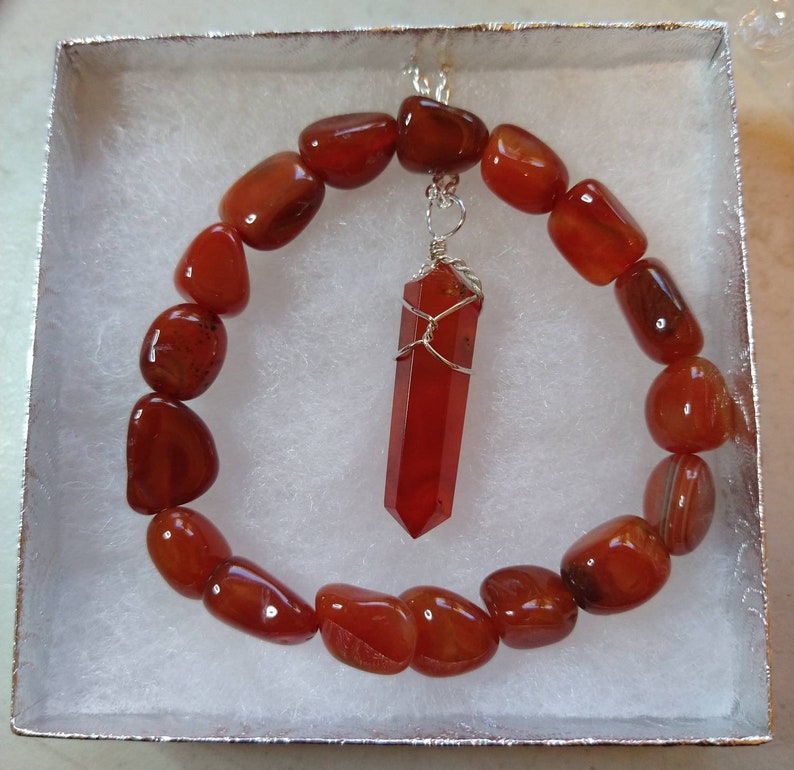 Op de afbeelding: Een carneliaan edelsteen armband en hanger set. De armband is gemaakt van gepolijste, rood-oranje stenen. De hanger is een bijpassende carneliaan kristal gewikkeld in zilverdraad. De set wordt gepresenteerd in een witte geschenkdoos.