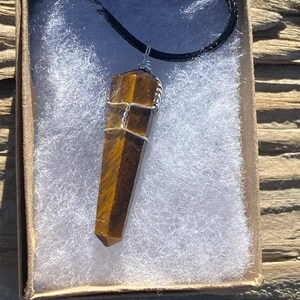 May include: A brown and gold tiger's eye crystal pendant wrapped in silver wire, hanging from a black cord, in a white and brown box.