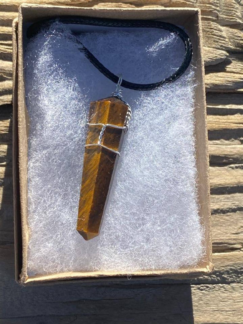 May include: A brown and gold tiger's eye crystal pendant wrapped in silver wire, hanging from a black cord, in a white and brown box.