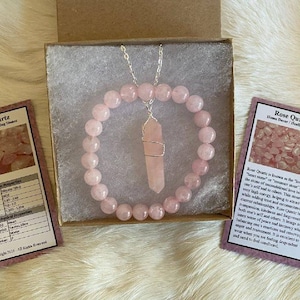 May include: A rose quartz jewelry set, including a beaded bracelet and a pendant necklace with a crystal point, presented in a gift box. Two cards with text about rose quartz are also included. The set is a soft pink color.