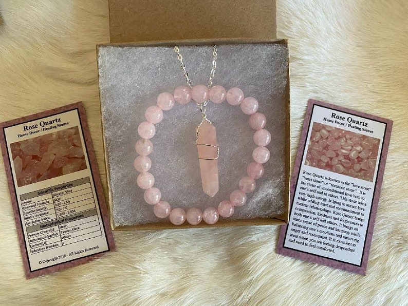May include: A rose quartz jewelry set, including a beaded bracelet and a pendant necklace with a crystal point, presented in a gift box. Two cards with text about rose quartz are also included. The set is a soft pink color.