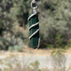 Pu&ograve; includere: Un ciondolo di cristallo verde scuro avvolto in filo d'argento, appeso a una catena d'argento. Il ciondolo ha una forma a punta ed &egrave; sospeso su uno sfondo sfocato di alberi e cielo.