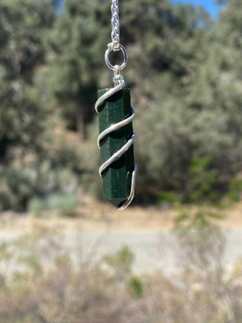 Pu&ograve; includere: Un ciondolo di cristallo verde scuro avvolto in filo d'argento, appeso a una catena d'argento. Il ciondolo ha una forma a punta ed &egrave; sospeso su uno sfondo sfocato di alberi e cielo.