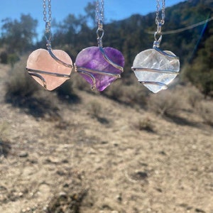 May include: Three silver heart-shaped pendants with wire wrapped around them. The pendants are made of rose quartz, amethyst, and clear quartz.