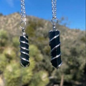 May include: Two black crystal pendants wrapped in silver wire on silver chains. The pendants are hanging against a blurred background of green foliage.