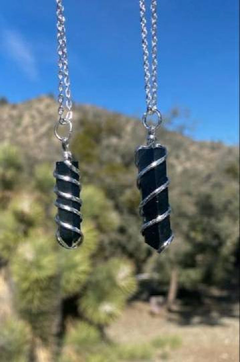 May include: Two black crystal pendants wrapped in silver wire on silver chains. The pendants are hanging against a blurred background of green foliage.