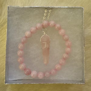 May include: A light pink beaded bracelet and necklace set. The bracelet and necklace feature round, light pink beads. The necklace has a pendant of a light pink crystal wrapped in silver wire. The jewelry is displayed in a white box.