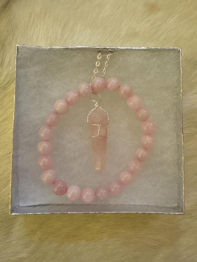 May include: A light pink beaded bracelet and necklace set. The bracelet and necklace feature round, light pink beads. The necklace has a pendant of a light pink crystal wrapped in silver wire. The jewelry is displayed in a white box.