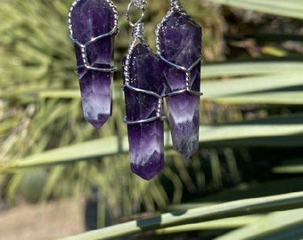 May include: Three amethyst crystal pendants with silver wire and chains. The crystals are a deep purple with white tips. The pendants are suspended against a blurred green backdrop.