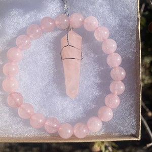May include: A rose quartz jewelry set in a gift box. The set includes a beaded bracelet and a pendant necklace with a wire-wrapped crystal point. The beads and crystal are a soft pink color.
