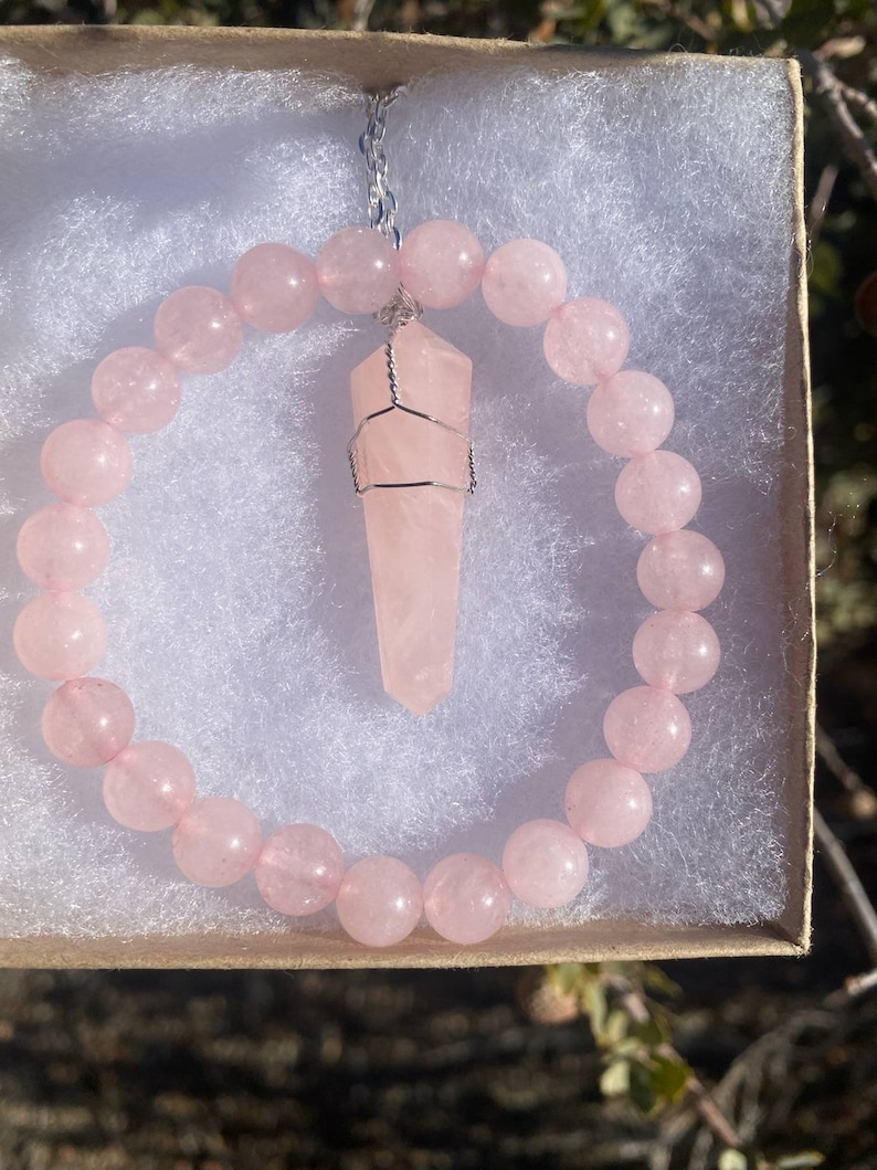 May include: A rose quartz jewelry set in a gift box. The set includes a beaded bracelet and a pendant necklace with a wire-wrapped crystal point. The beads and crystal are a soft pink color.