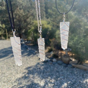 May include: Three clear crystal pendants with silver wire wrapped around them, hanging from different chains. The pendants are hanging in front of a blurred background of trees and rocks.