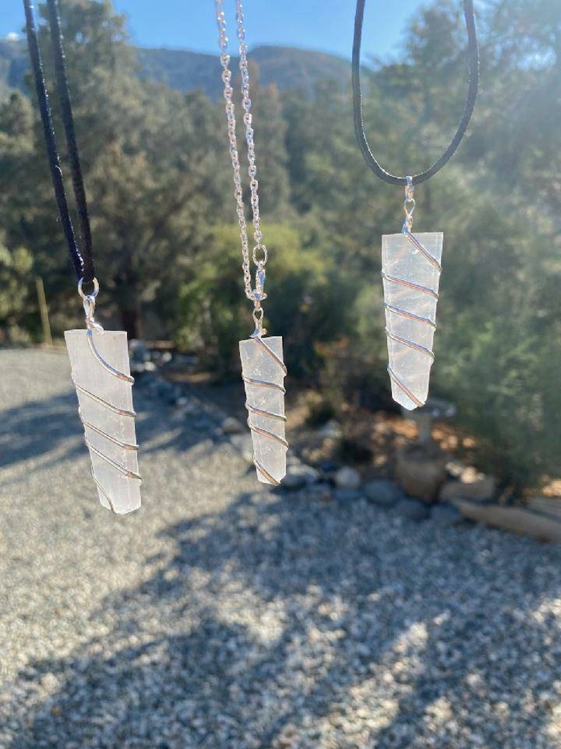 May include: Three clear crystal pendants with silver wire wrapped around them, hanging from different chains. The pendants are hanging in front of a blurred background of trees and rocks.