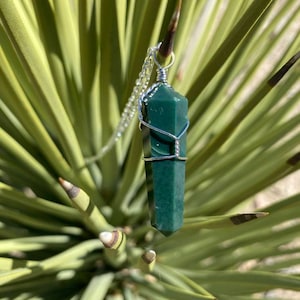 Pu&ograve; includere: Un ciondolo di cristallo verde scuro avvolto in filo d'argento, appeso a una catena d'argento. Il ciondolo &egrave; a fuoco, mentre lo sfondo &egrave; sfocato.