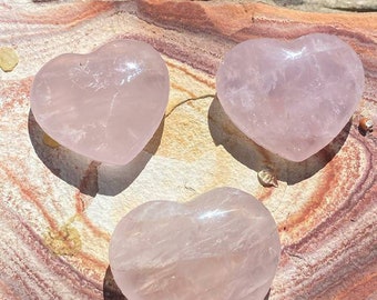 May include: Three pink rose quartz heart shaped stones on a brown and white striped surface.