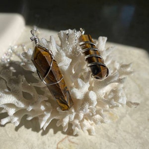 May include: Two gold tiger's eye crystal pendants wrapped in silver wire, hanging from silver chains. The pendants are displayed on a white coral-like surface.