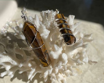 May include: Two gold tiger's eye crystal pendants wrapped in silver wire, hanging from silver chains. The pendants are displayed on a white coral-like surface.