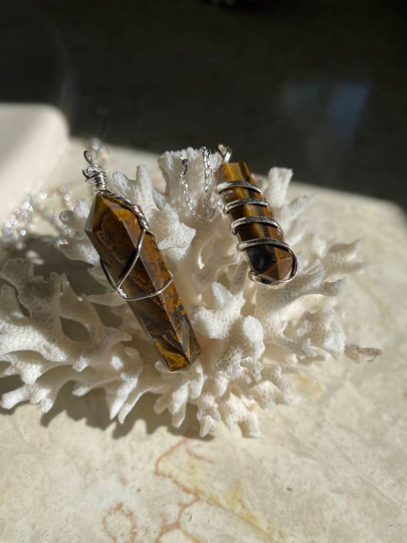 May include: Two gold tiger's eye crystal pendants wrapped in silver wire, hanging from silver chains. The pendants are displayed on a white coral-like surface.