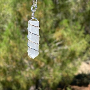 May include: A white selenite crystal wrapped in silver wire, hanging from a silver chain. The crystal is a long, thin, rectangular shape with a pointed end. The background is blurred and green.