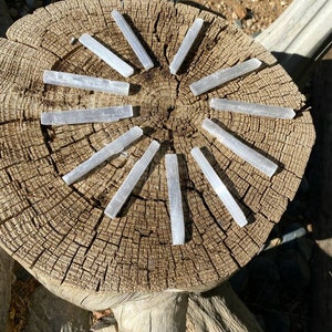 May include: Twelve elongated, translucent white selenite crystal wands arranged in a circular pattern. Each crystal features a fibrous, striated texture with a natural, raw appearance. The wands are positioned radially on a circular wooden log slice.