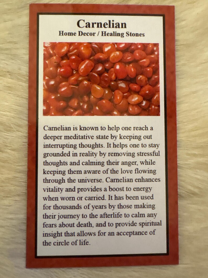 Op de afbeelding: Een kaart met het woord "Carnelian" en de tekst "Home Decor / Healing Stones". De afbeelding toont een close-up van talrijke gepolijste, rood-oranje carnelian stenen. De kaart bevat ook tekst over de eigenschappen van de steen.