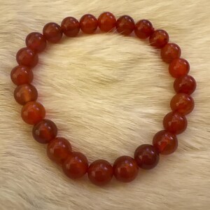 Op de afbeelding: Een armband gemaakt van gepolijste, ronde, rood-oranje kralen. De kralen zijn aan elkaar geregen en vormen een cirkelvorm. De armband wordt gepresenteerd op een zacht, wit, harig oppervlak.