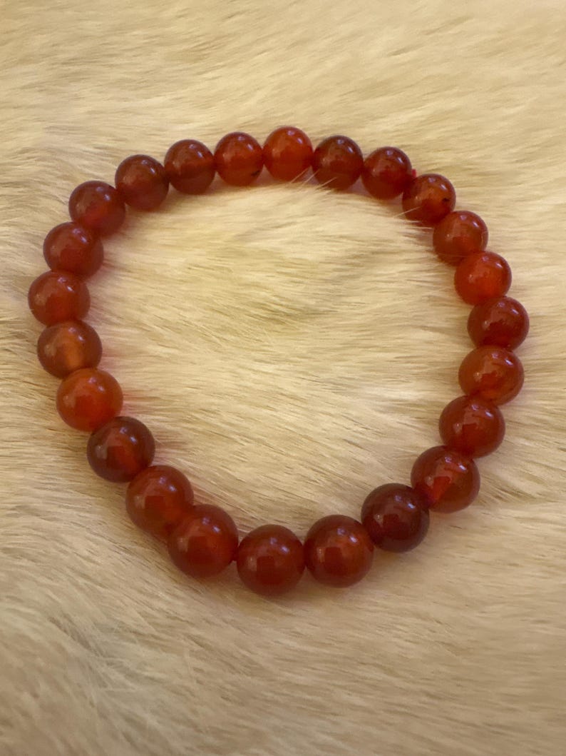 Op de afbeelding: Een armband gemaakt van gepolijste, ronde, rood-oranje kralen. De kralen zijn aan elkaar geregen en vormen een cirkelvorm. De armband wordt gepresenteerd op een zacht, wit, harig oppervlak.