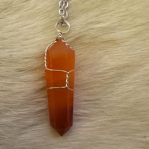 Op de afbeelding: Een hangerketting met een gefacetteerd, oranjerood kristal, gewikkeld in zilverkleurig draad. Het kristal hangt aan een zilveren ketting. De ketting wordt gepresenteerd tegen een zachte, lichtgekleurde achtergrond.