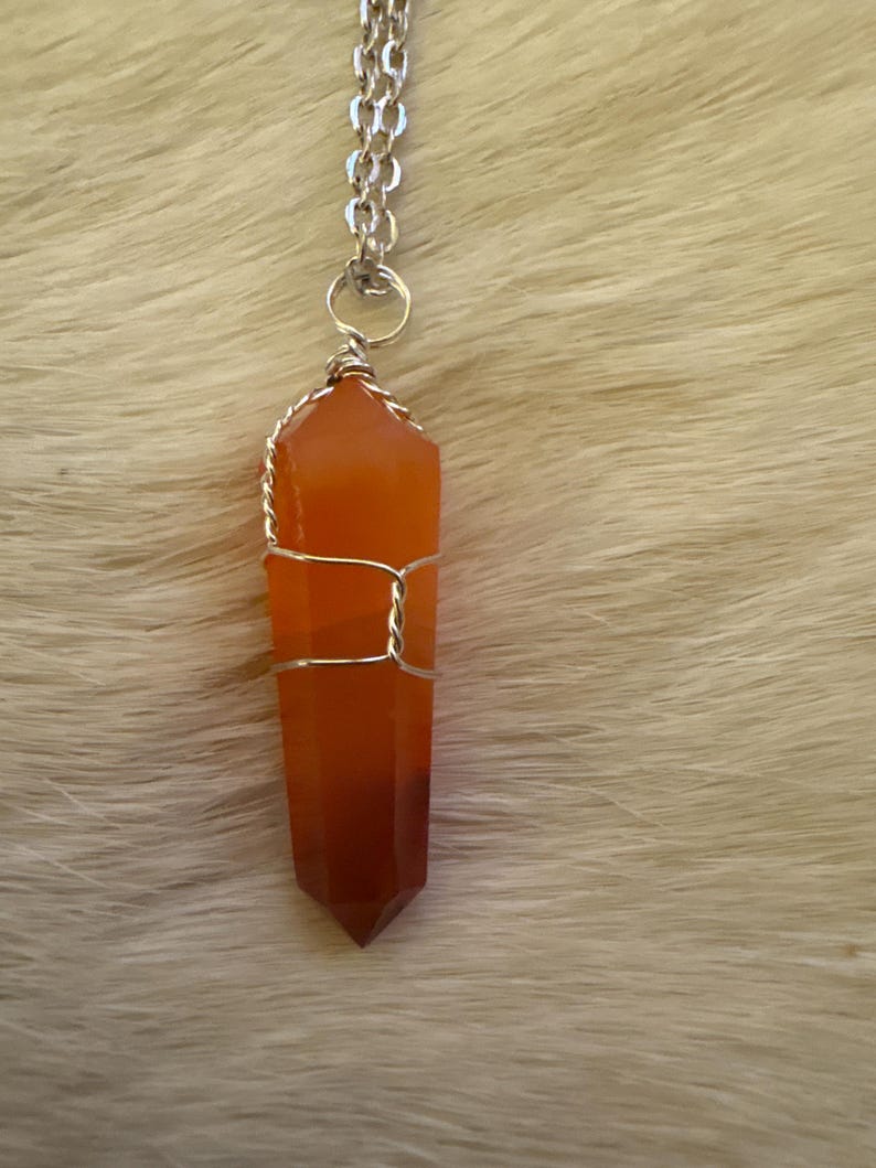 Op de afbeelding: Een hangerketting met een gefacetteerd, oranjerood kristal, gewikkeld in zilverkleurig draad. Het kristal hangt aan een zilveren ketting. De ketting wordt gepresenteerd tegen een zachte, lichtgekleurde achtergrond.