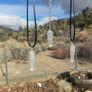May include: Three silver wire wrapped clear crystal point pendants on black cords. The pendants are hanging against a backdrop of a cloudy sky and a distant mountain range.