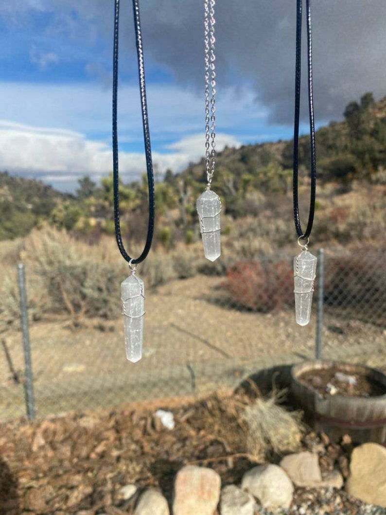 May include: Three silver wire wrapped clear crystal point pendants on black cords. The pendants are hanging against a backdrop of a cloudy sky and a distant mountain range.