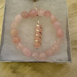 May include: A pink rose quartz bracelet and pendant necklace set. The bracelet is made of rounded pink stones. The pendant is a pink rose quartz crystal wrapped in silver wire, suspended from a silver chain. The jewelry is presented in a silver box.